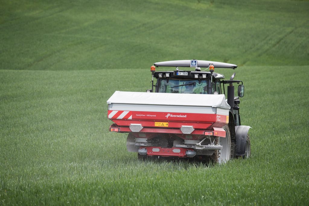 Traktor med kunstgjødselspreder arbeider på en grønn kornåker. Foto.