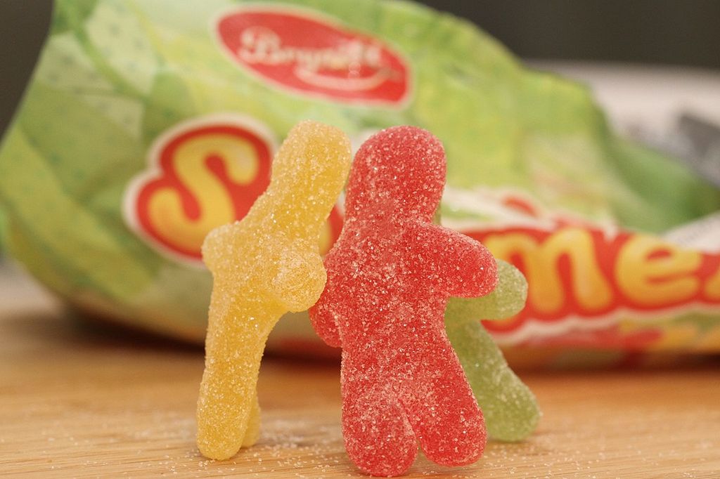 Tre sukrete gelefigurer, en gul, en rød og en grønn, står oppreist på ei bordplate og "holder" i hverandre. I bakgrunnen er emballasjen til seigmennene med logoen til Brynild synlig. Foto.