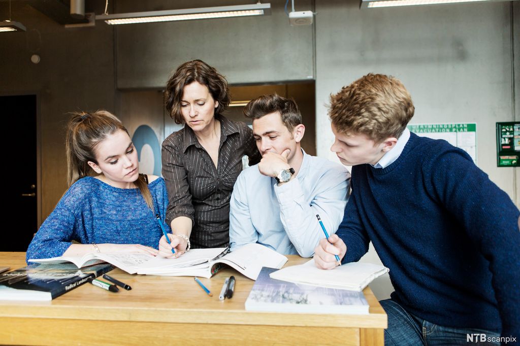 Ein lærar rettleier tre studentar i eit klasserom. Dei har oppslåtte skrivebøker, lesebøker og blyantar og pennar på eit bord som dei står og sit rundt. Foto.