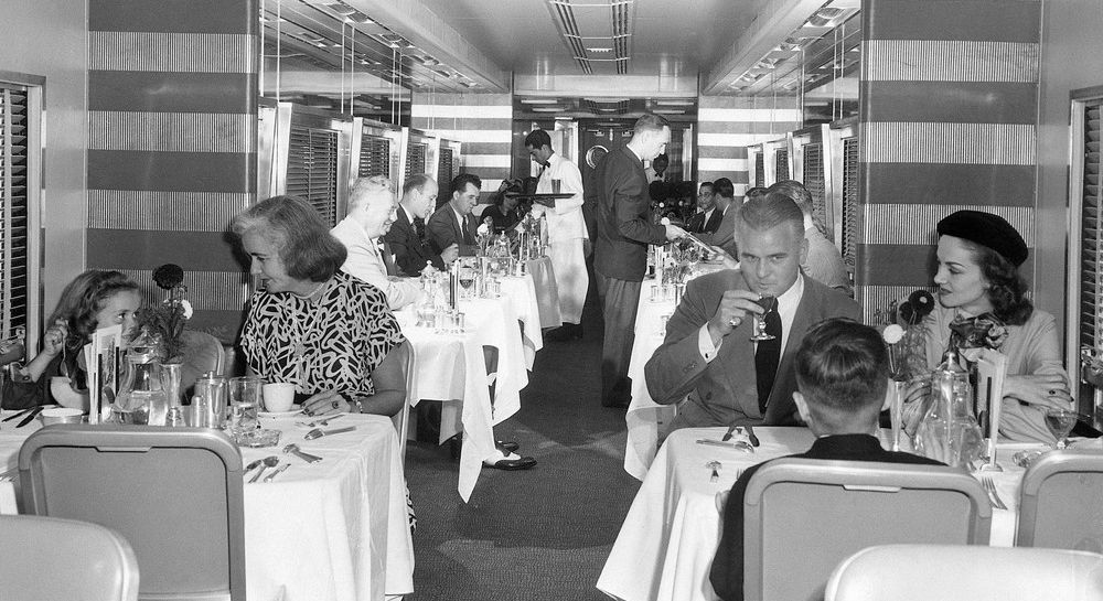 Restaurantvogn på toget. Bordene har hvite duker, og menneskene er pent kledde i dress og kjoler. Foto i svarthvitt. 