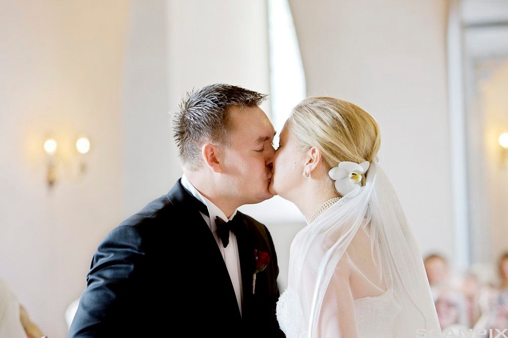 En brudgom i mørk dress kysser en brud kledd i hvitt. Foto.