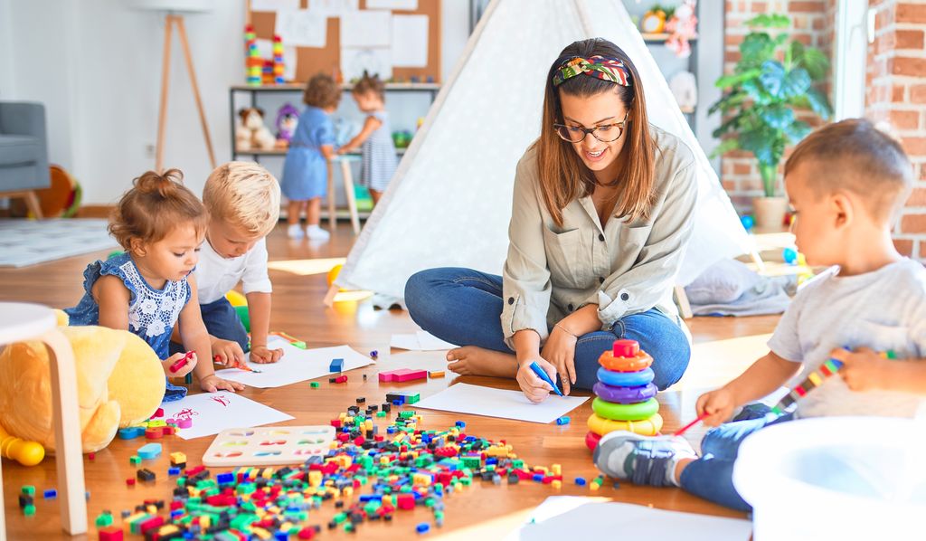 Ung kvinnelig ansatt sitter på gulvet med ei gruppe barn. Hun tegner på et ark. To små barn tegner på egne ark. En liten gutt ser på det kvinnen tegner mens han bygger med fargerike klosser som også ligger ut over gulvet. I bakgrunnen står to jenter og leker med noe ved veggen. Foto.