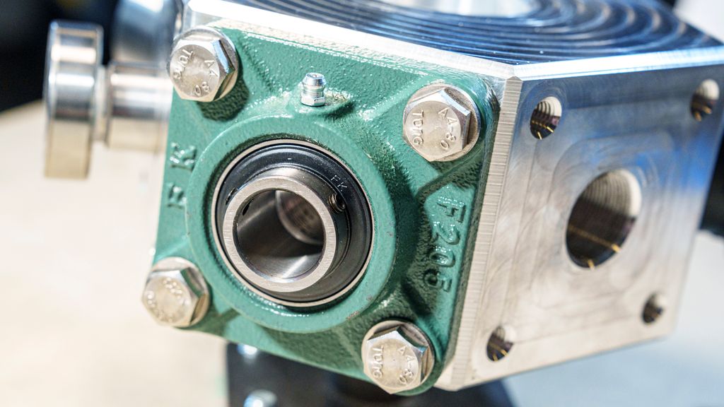 En grønn metallkonstruksjon med et sirkulært hull som er festa til en større enhet med fire skruer i hjørnene. Foto.