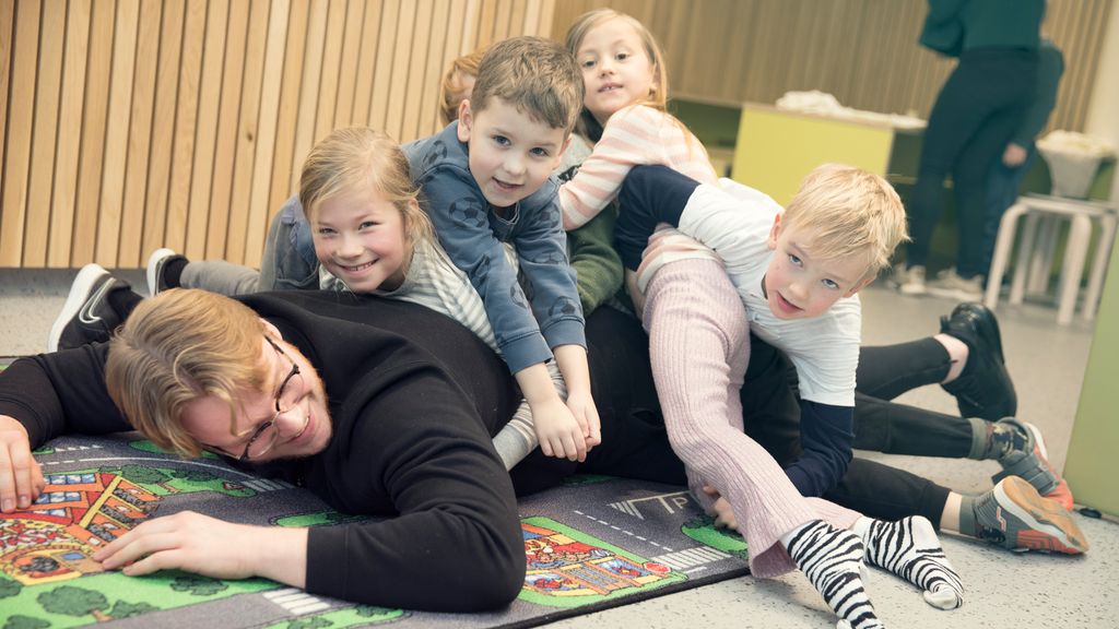 Fem barn leker med en voksen mann på gulvet. Barna ligger i en stor haug oppå ryggen til mannen. Foto.