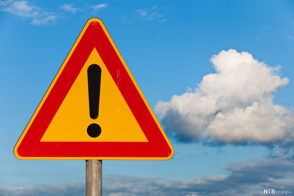 Et trekantet skilt med et svart utropstegn i midten og rød kant rundt trekanten. I bakgrunnen er små skyer på en blå himmel. Foto.
