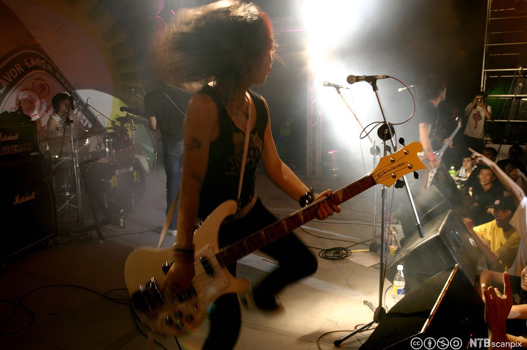 Person med flagrende hår spiller på bassgitar på en scene. Foto.