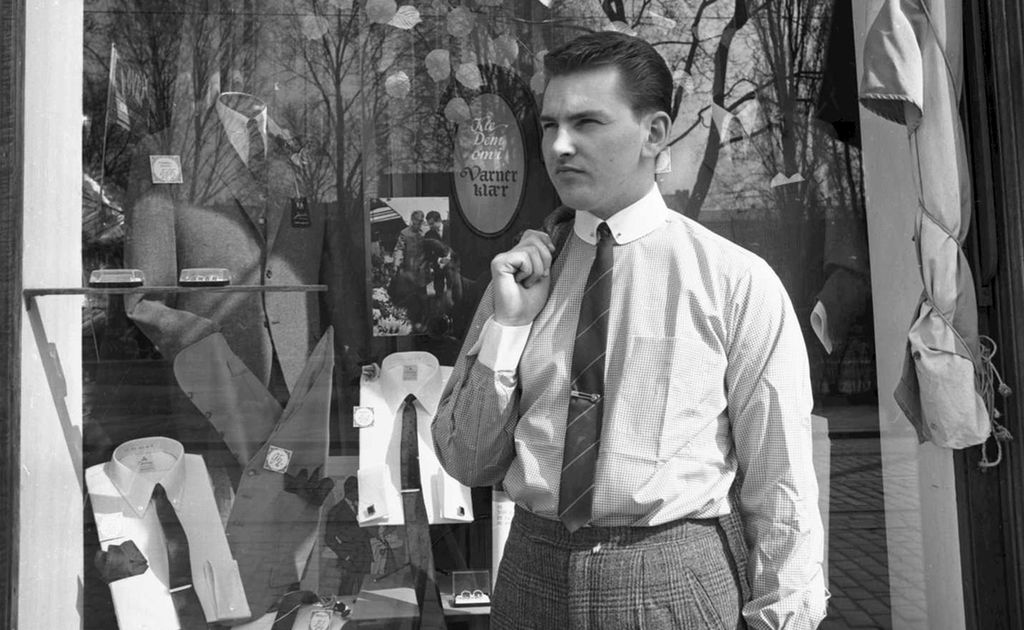 Frank Varner utenfor butikken sin på Grünerløkka i Oslo. Han er kledd i rutete dressbukse, rutete skjorte og slips med slipsnål. Han holder dressjakka i høyre hånd over skulderen. Håret hans er finkjemmet bakover. Foto.