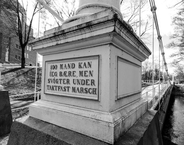 Gammelt advarselsskilt på ei hengebru med teksten 100 mand kan jeg bære, men svigter under taktfast marsch. Svart-hvitt foto.