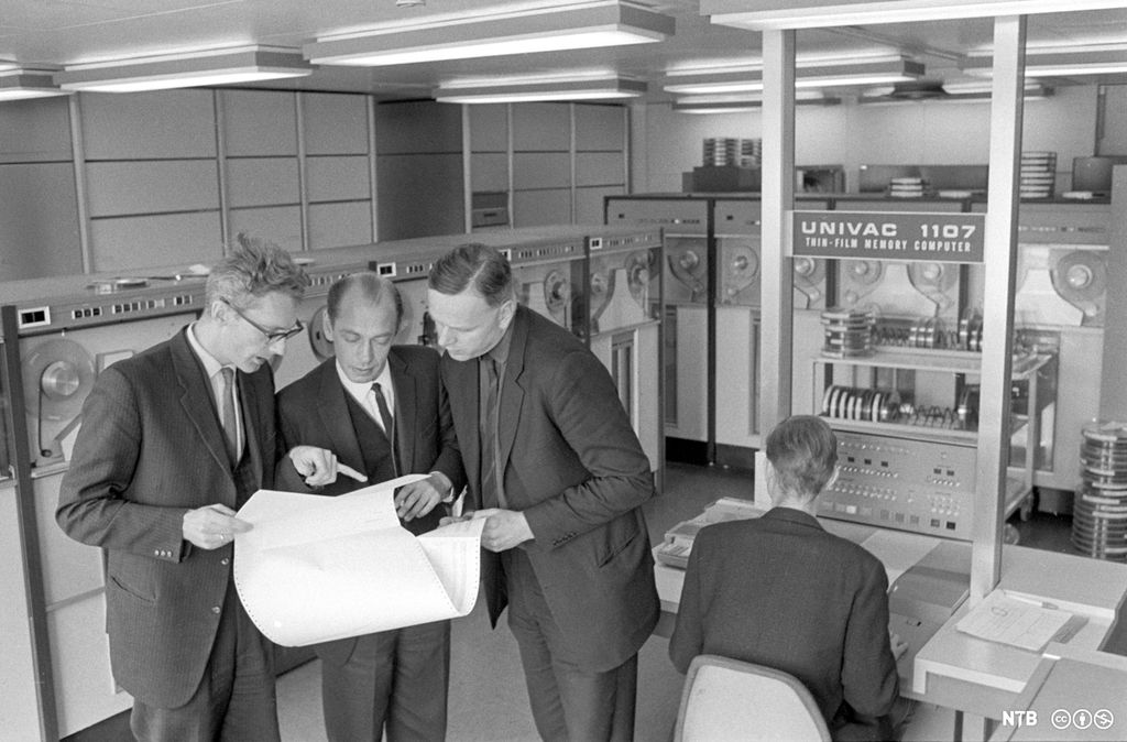 Tre menn i dress studerer eit ark med utskrift av programmeringskode. Ein fjerde mann sit med ryggen til ved ein datamaskinterminal av typen "Univac 1107". Foto.