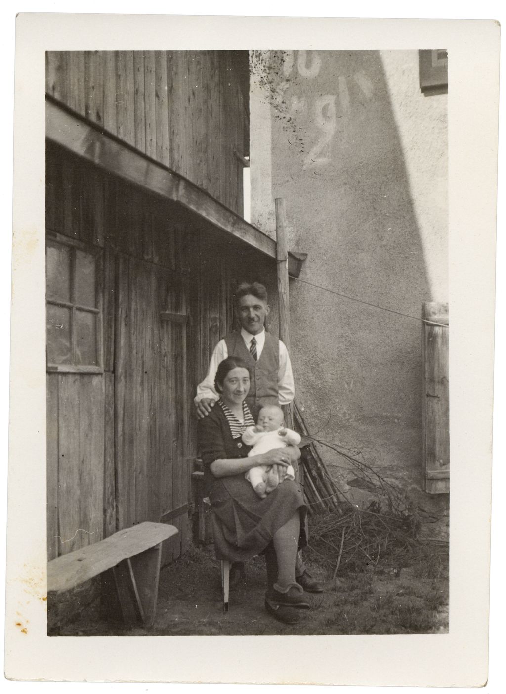 Ein mann og ei kvinne i slutten av tjueåra. Kvinna sit på ein stol og held eit spedbarn i armane, mannen står bak ho og har lagt den eine handa si på skuldra hennar. Begge har på seg enkelt pentøy. I bakgrunnen delar av ein låve og av eit murhus. Gammalt foto.