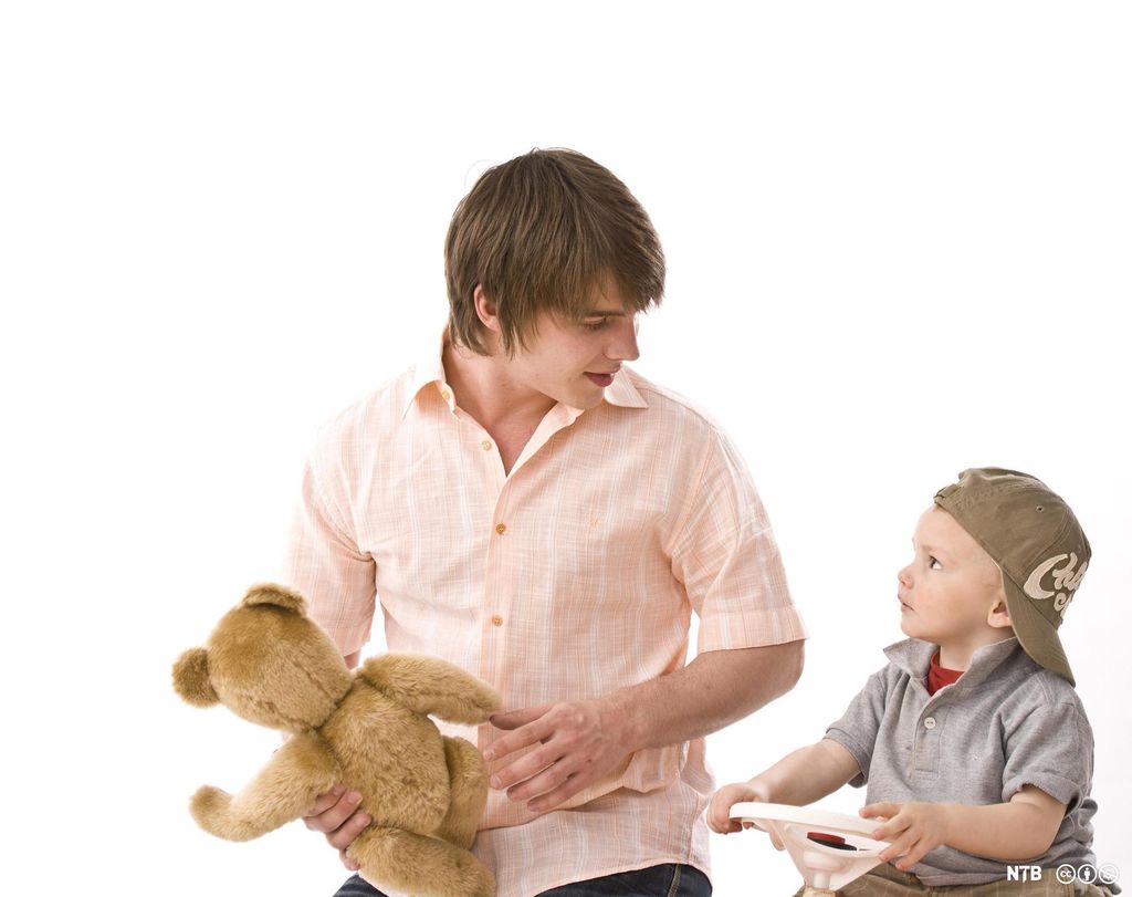 En ung mann leker sammen med en liten gutt. Mannen holder en bamse og gutten holder i rattet på en liten lekebil. De ser på hverandre. Foto.