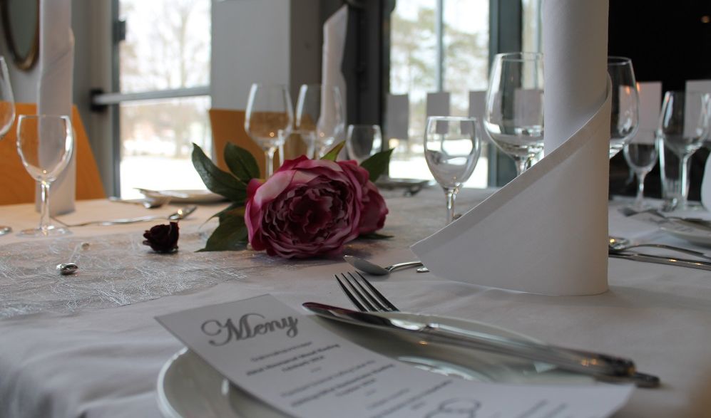 Pent dekt bord med kvit duk, vinglas, bestikk, tøyserviettar, menykort og blomstrar som pynt. Foto.