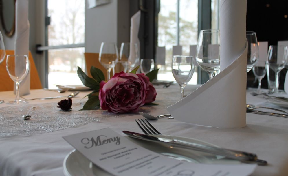 Pent dekket bord med hvit duk, vinglass, bestikk, tøyservietter, menykort og blomster som pynt. Foto.
