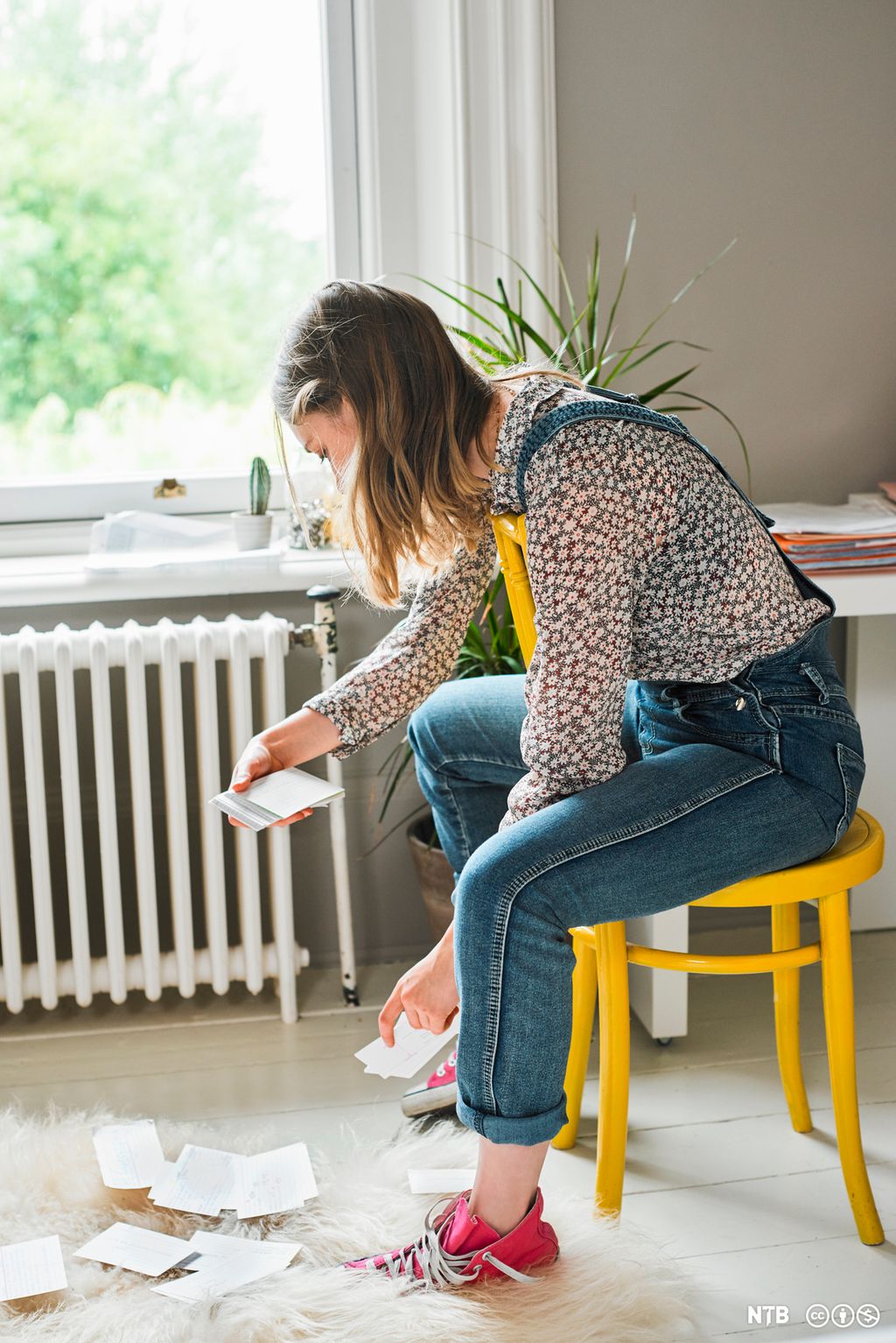 Ung, kvinnelig student sitter på en gul stol og studerer små notatark som hun har lagt på gulvet. Foto.