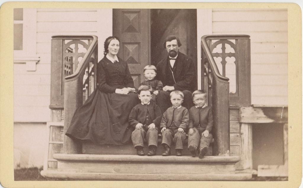 En familie sitter på trappa til et hus: mor i svart kjole, far i svart dress, tre små gutter i dress og ei lita jente i kjole. Foto.