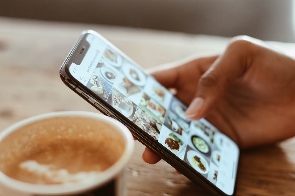 Nærbilete av ein kaffikopp og ei hand som held ein mobiltelefon og blar på Instagram. Foto.