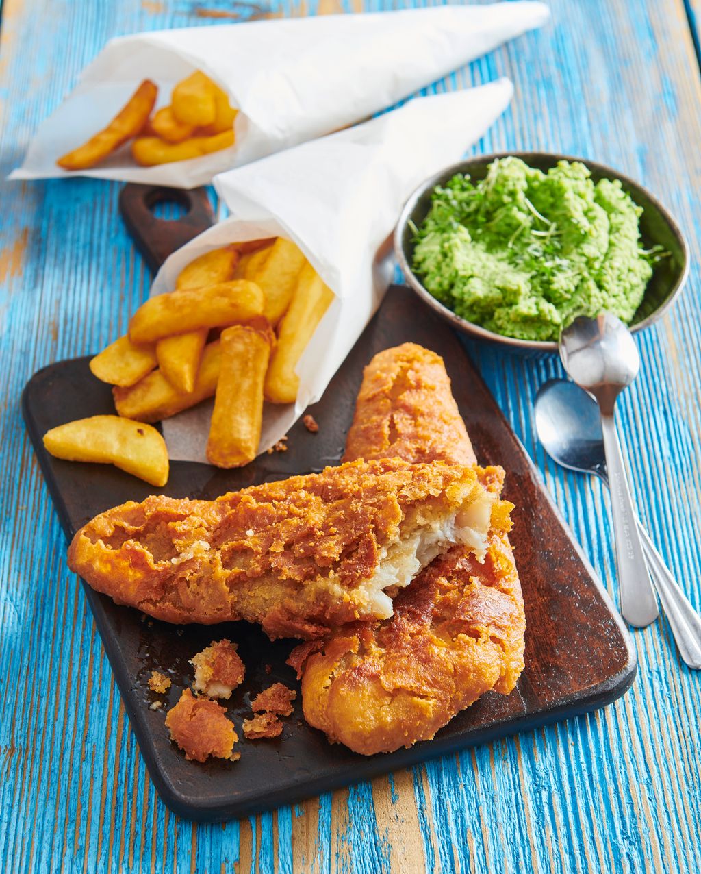Fish and chips med ertestuing. Fisken er anrettet på ei trefjøl og pommes fritesen er i et papirkremmerhus. Foto.