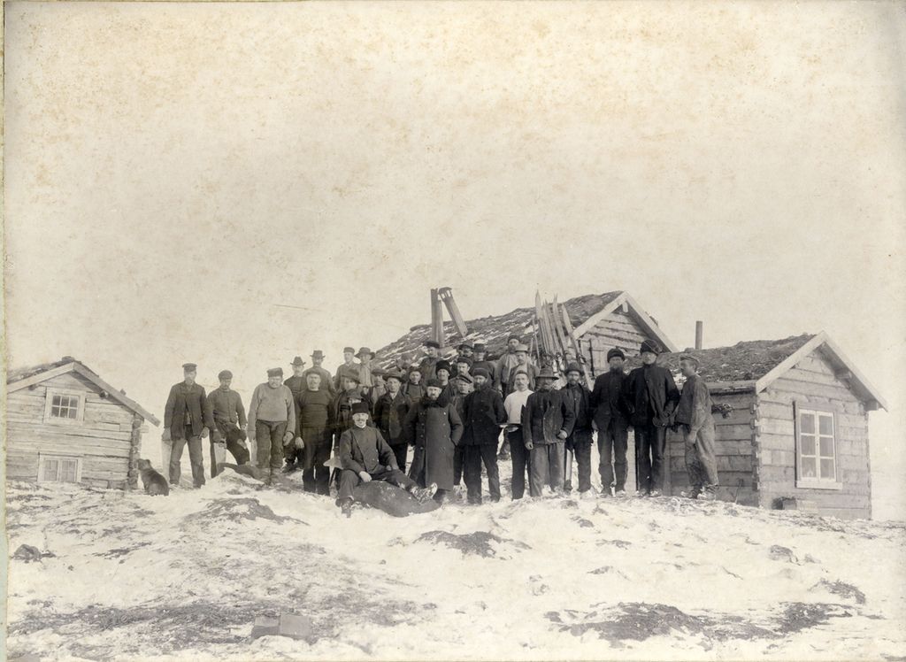 Arbeidere foran den eldste stollen (gruvegangen) på kobberverket i Kåfjord. Det er snø på bakken og små laftede trehus rundt der de står. Sort/hvitt-foto.