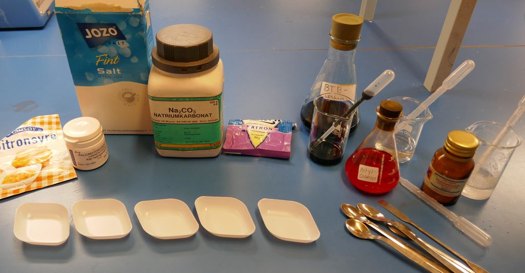 Kjemikaler, begerglass, beger og spatel på en benk. Foto.