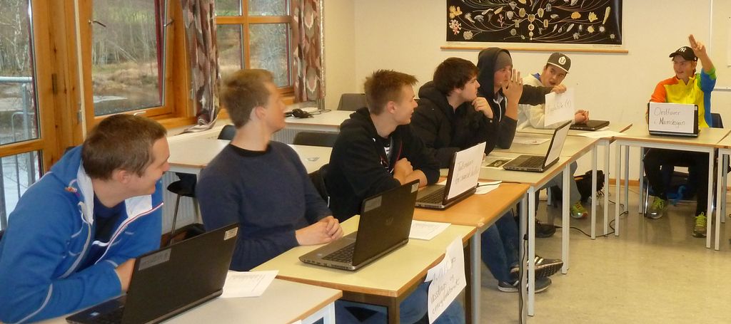I et klasserom er pultene ordnet i en hestesko. Foran hver elev er et stort ark der det står hvilken rolle eleven spiller i debatten. Foto.