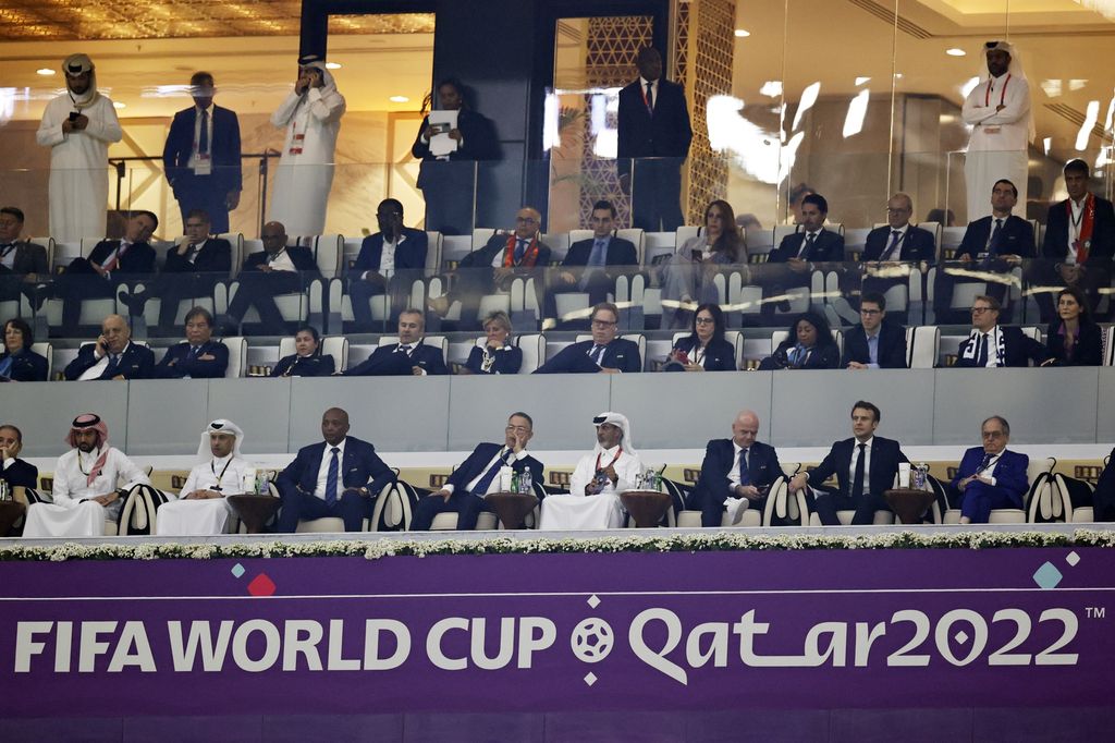 Vestlig kledde og arabisk-kledde tilskuere på en tribune. Under tribunen står et skilt med Fifa World Cup Qatar 2022. Foto. 