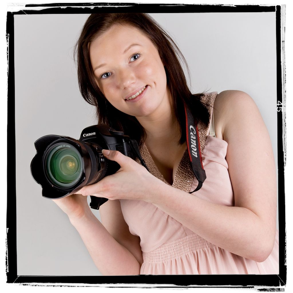 Jente i rosa kjole holder en hånd under og en hånd på siden av et speilreflekskamera. Foto.