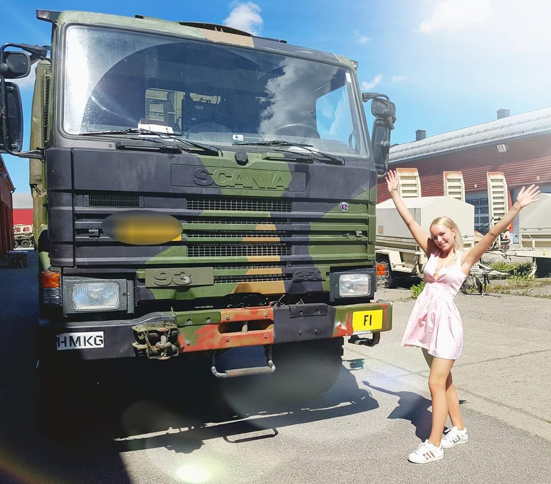 Mathilde Grøndalen Lund står ved siden av en stor lastebil. Hun er kledd i rosa kjole og hvite sneakers, og hun holder hendene i været og smiler. Foto.