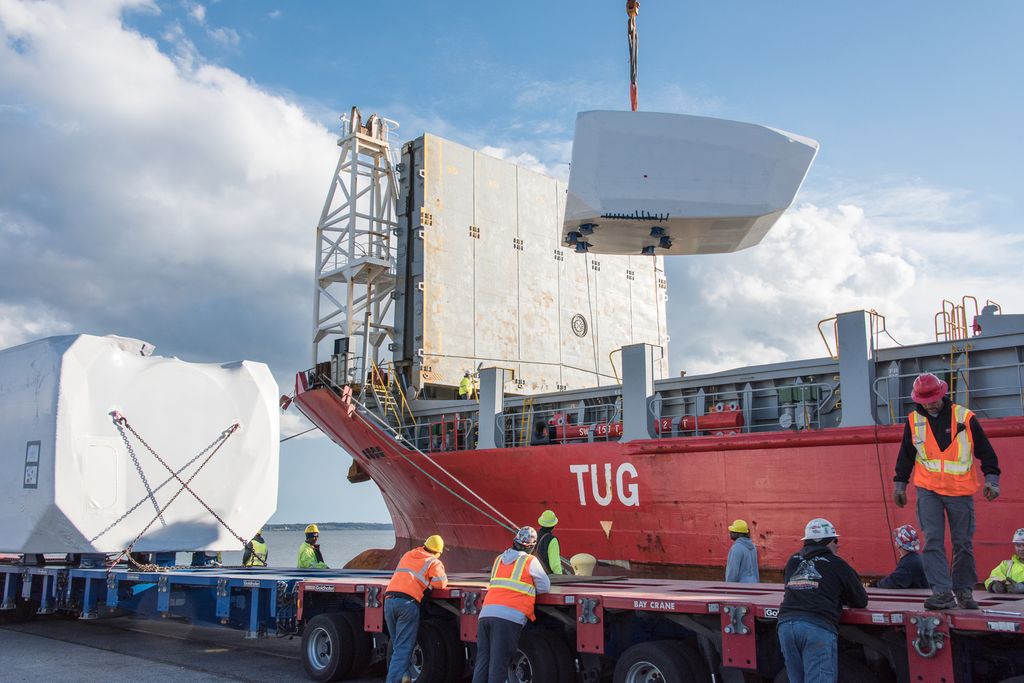 En stor maskindel løftes med kran mellom lasteplan og skip. Mange arbeidsfolk med hjelm og reflekterende arbeidstøy følger med. Foto.