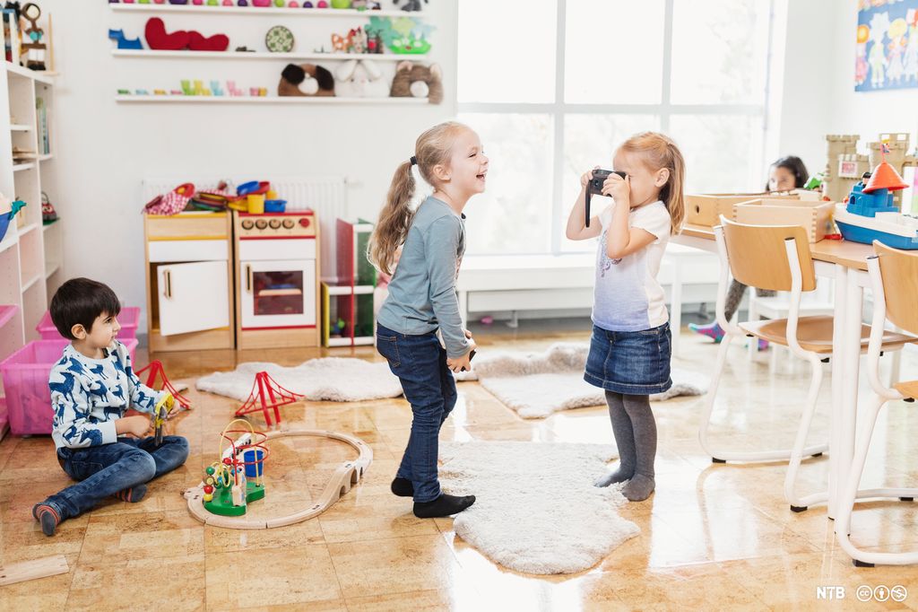 Ein gut sit på golvet og leiker med ein togbane. To jenter leier at den eine fotograferer den andre. Det er fleire leiker i rommet og til høgre er det bord og stolar. Foto.