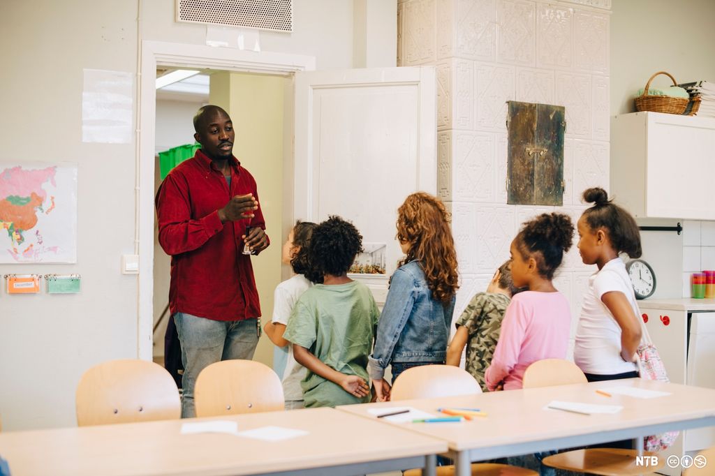 Seks barn står stilt opp foran læreren sin ved døra i et klasserom. Barna er alle ulike. Foto.