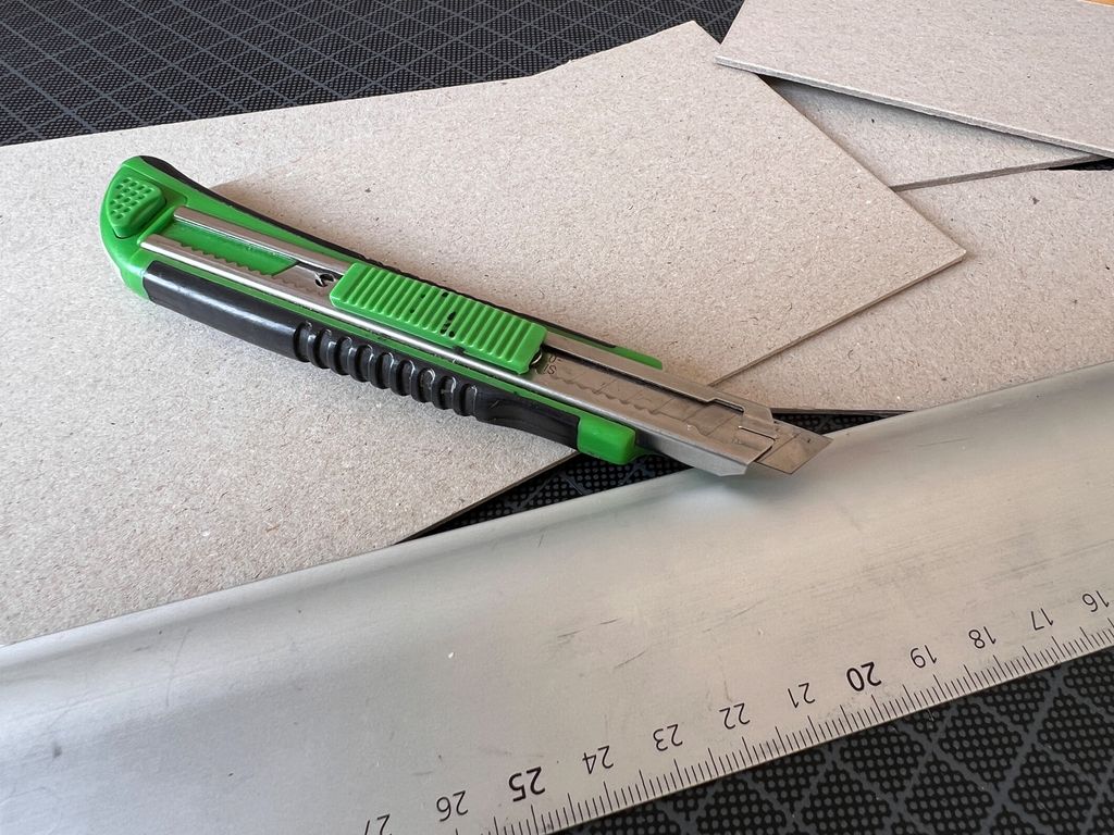 Tapetkniv og linjal av metall som ligger på biter av bokpapp. Foto.