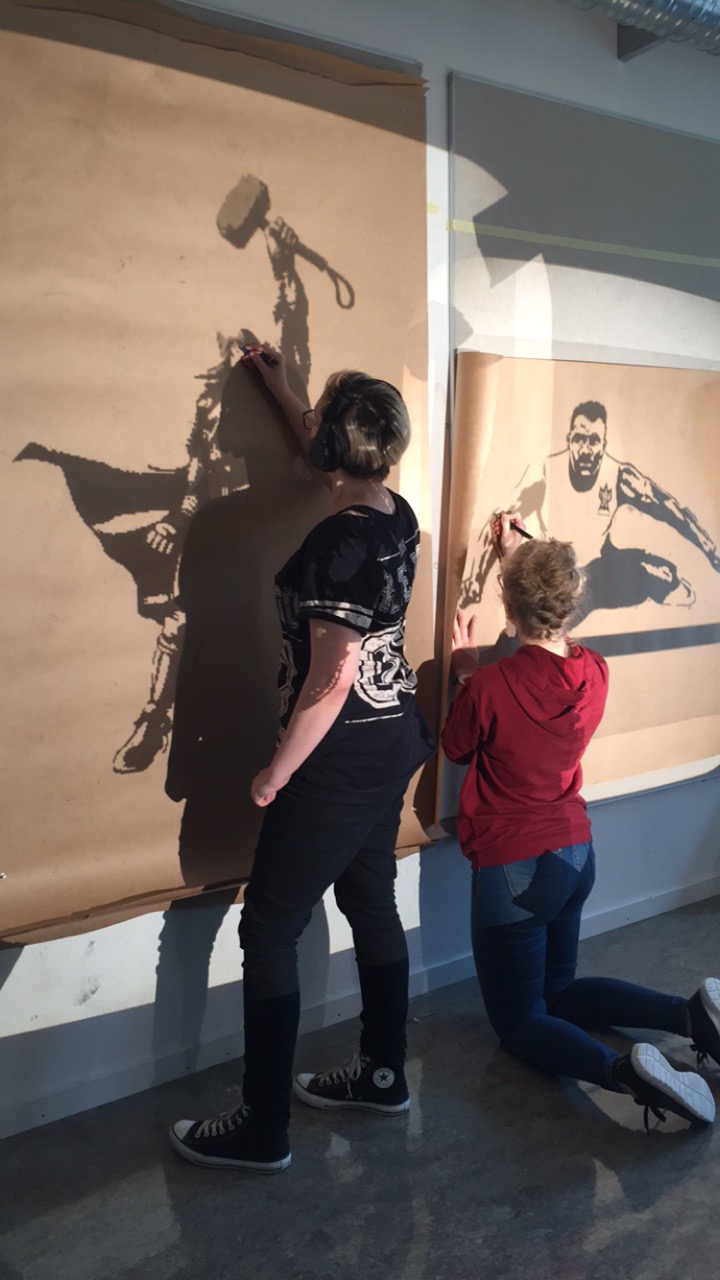 To elever tegner på gråpapir montert på en vegg i lyset fra en overheadprojektor. Foto.