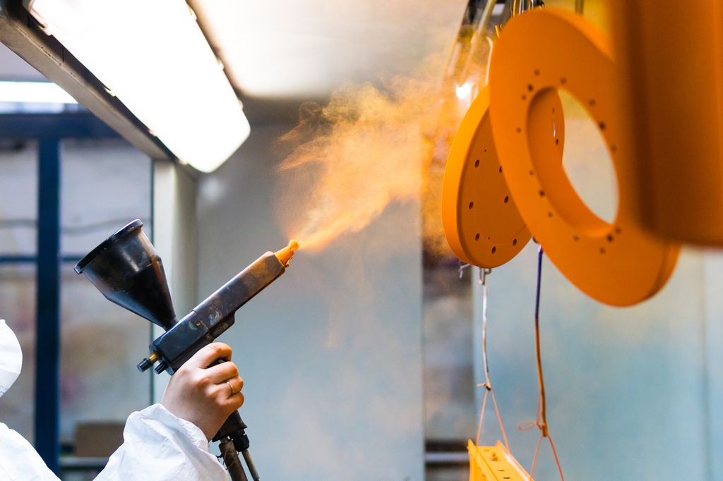 Person med verneutstyr pulverlakkerer metalldeler. Foto.