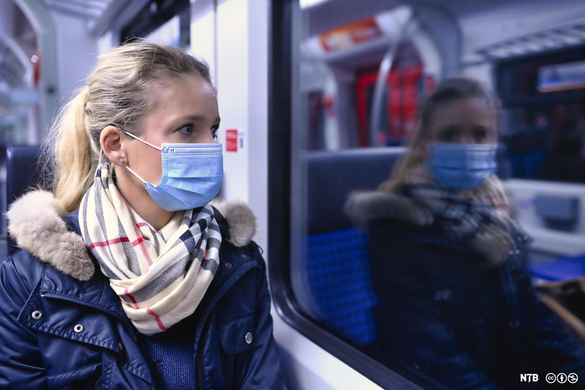 Kvinne med ansiktsmaske sit på eit tog og ser ut vindauget. Foto.
