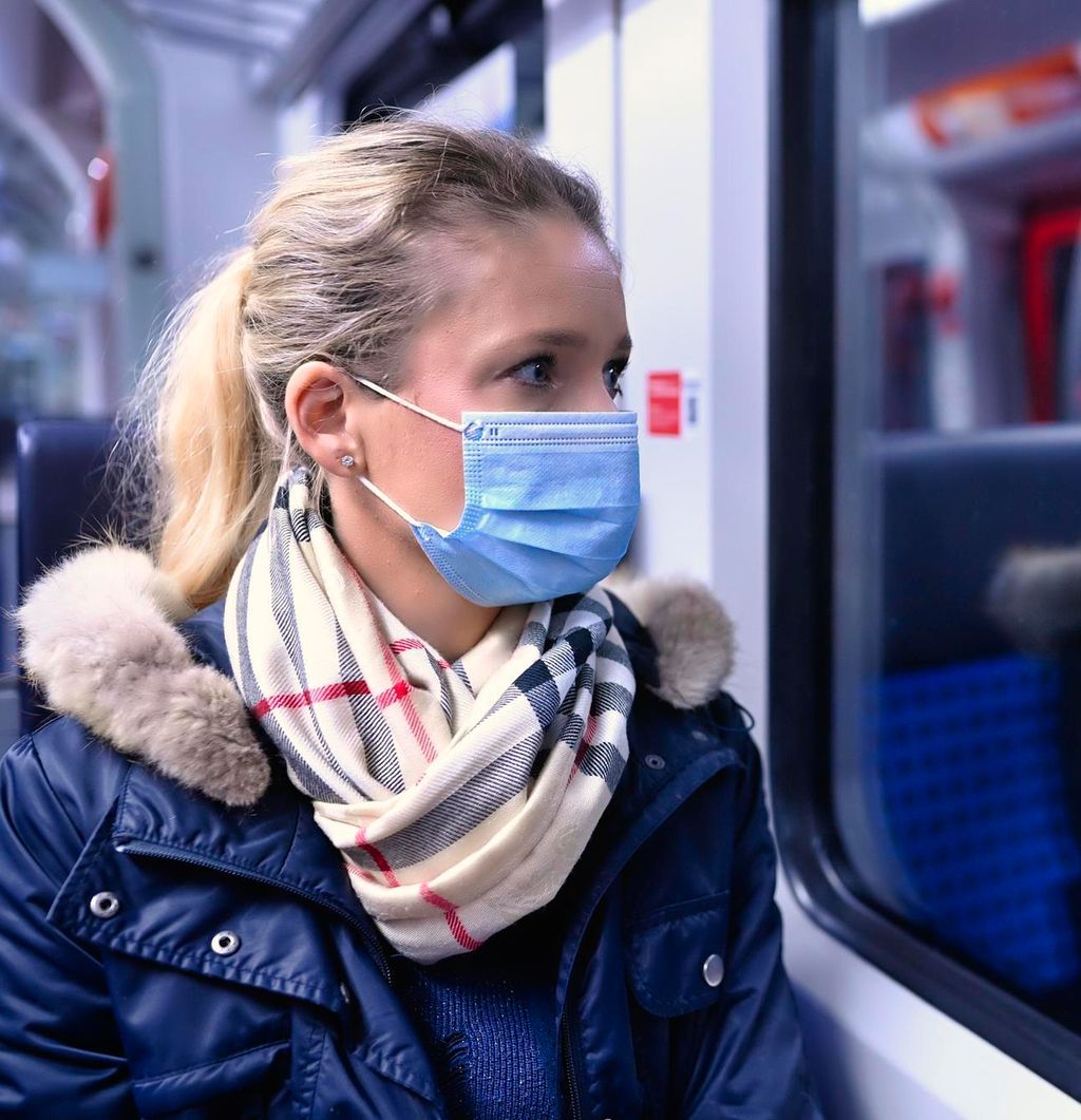 Kvinne med ansiktsmaske sitter på et tog og ser ut vinduet. Foto.
