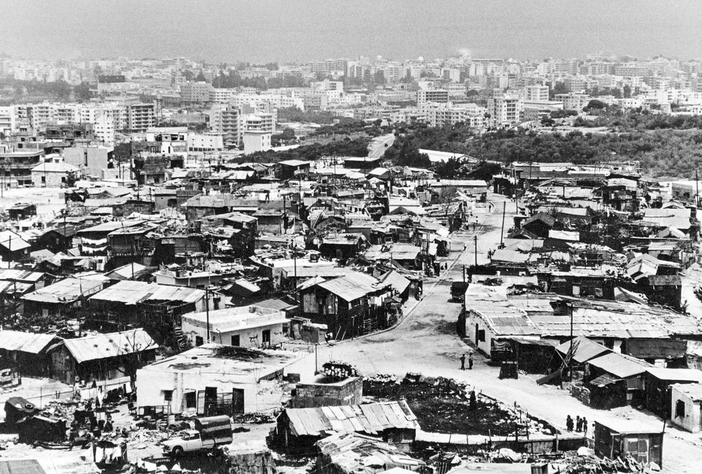 Leiren består av einetasjes enkle hus. I bakgrunnen er det ein moderne by. Foto.