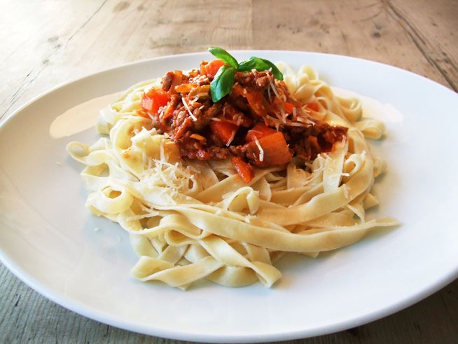 Pasta bolognese anrettet på en hvit tallerken. Revet ost og et grønt blad er brukt som garnityr. Foto.