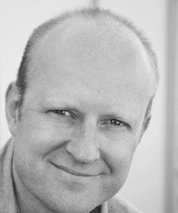 Nærbilde av ansiktet til en glattbarbert og smilende mann. Svart-hvitt foto.