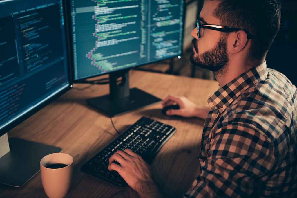 Mann sitter foran en datamaskin med to skjermer. Skjermene er fylt med linjer med kode. Foto.