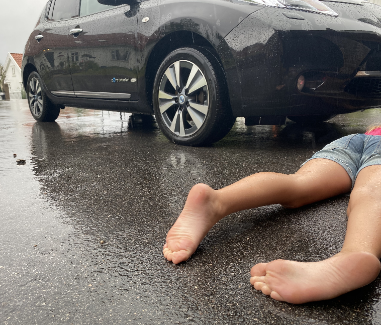 Ei jente ligger på magen på asfalten foran en bil, overkroppen er utenfor bildet. Beina ligger litt vridd. Hun er kledd i shorts, men det regner. Foto.
