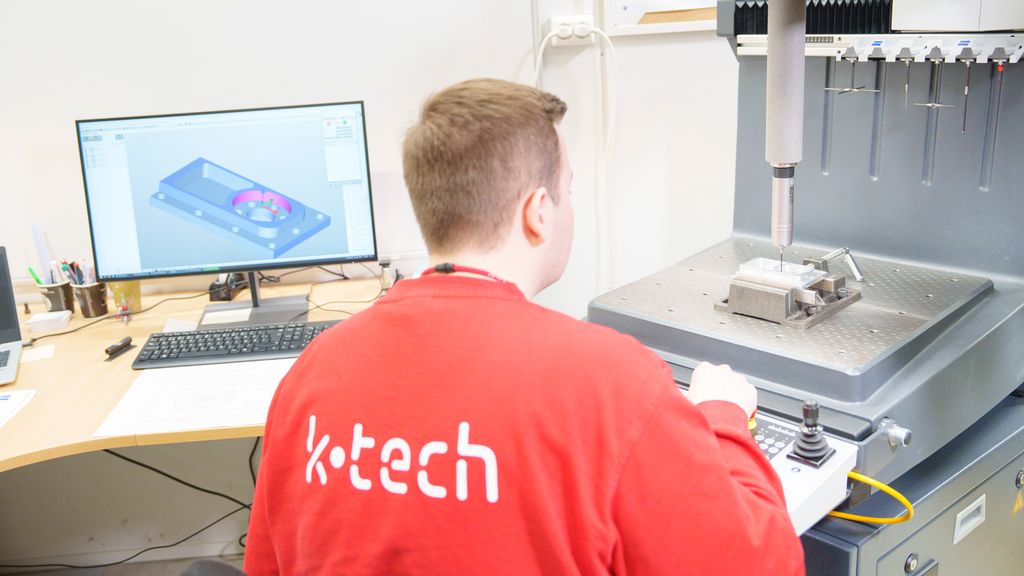 Del festa i en maskinstikke på ei planskive. Over delen henger det en målespiss. I bakgrunnen vises en 3D-modell av delen på en dataskjerm. Foto.