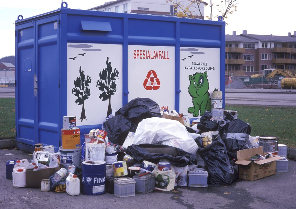 Sekker med søppel, bilbatterier, kanner og malingsspann står stablet på utsida av en stor blå konteiner med påskriften spesialavfall. Foto.