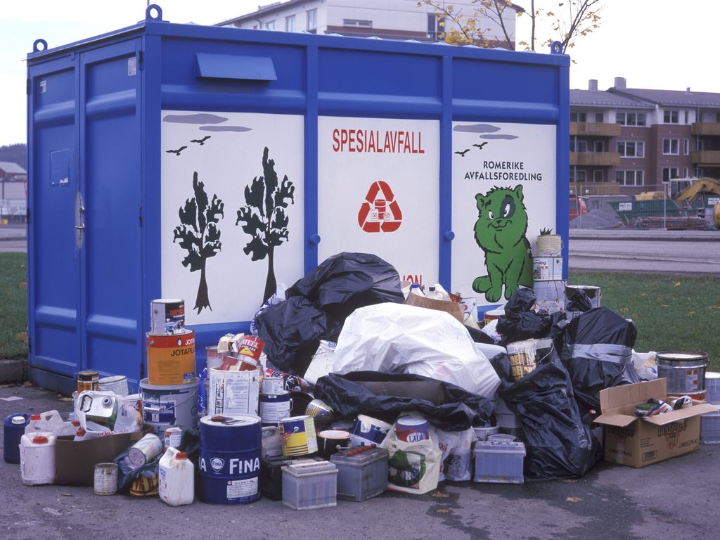 Sekker med søppel, bilbatteri, kanner og spann står stabla på utsida av ein stor blå konteinar med påskrifta SPESIALAVFALL. Foto.