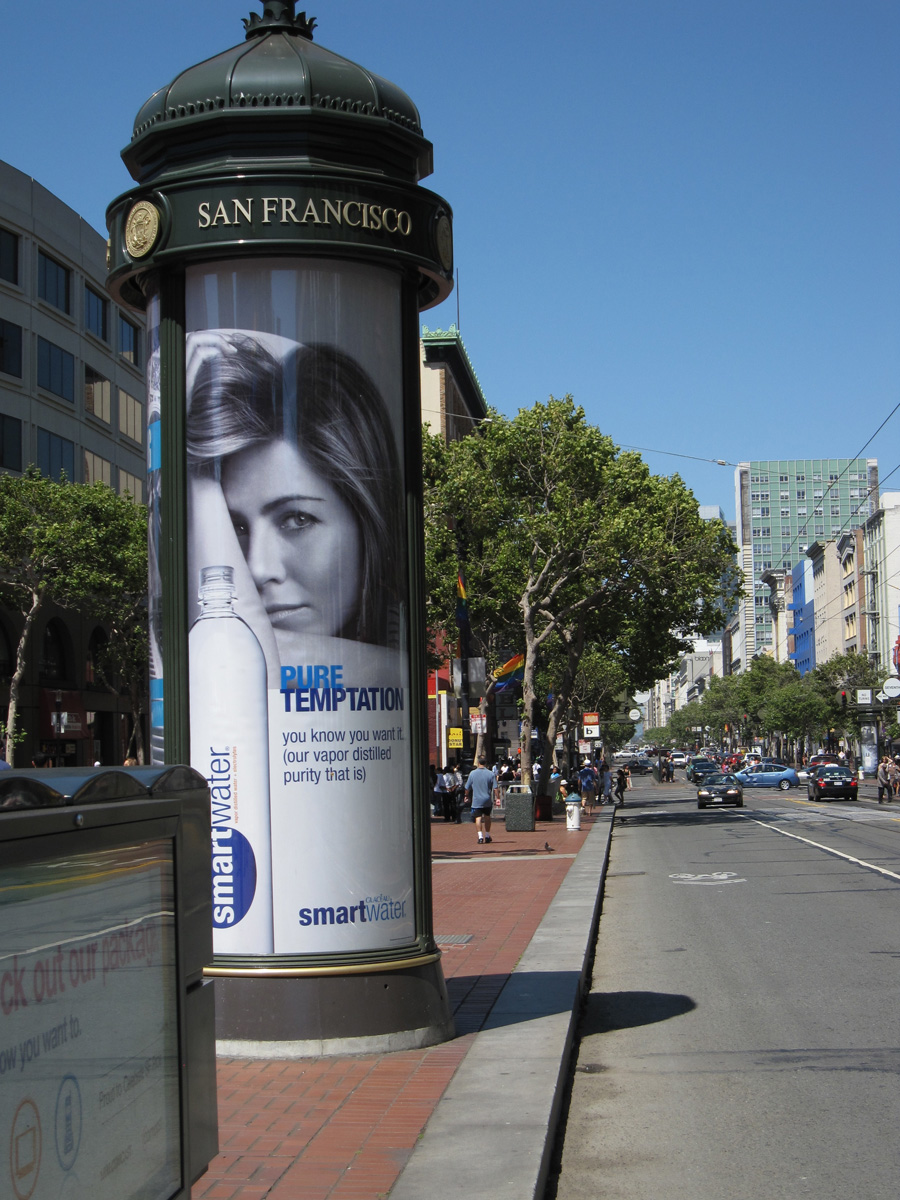 Et urbant miljø der det er montert en reklameplakat på en stolpe langs en trafikkert vei. Plakaten reklamerer for flaskevannet smartwater og viser en kvinne og ei flaske fra produsenten, i tillegg til slagord for produktet, pure temptation. Foto.