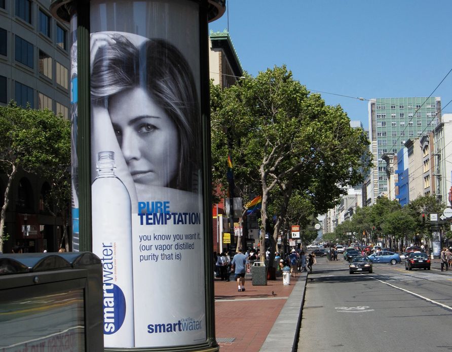 Et urbant miljø der det er montert en reklameplakat på en stolpe langs en trafikkert vei. Plakaten reklamerer for flaskevannet smartwater og viser en kvinne og ei flaske fra produsenten, i tillegg til slagord for produktet. Foto.