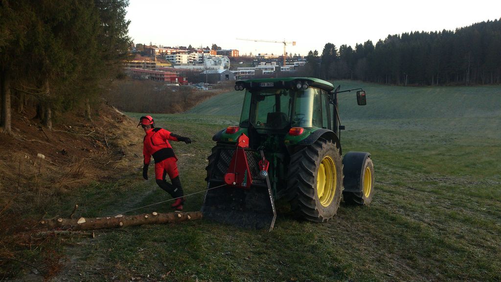 En traktor med påmontert vinsj trekker en tømmerstokk. En person med verneutstyr står ved siden av. Foto.