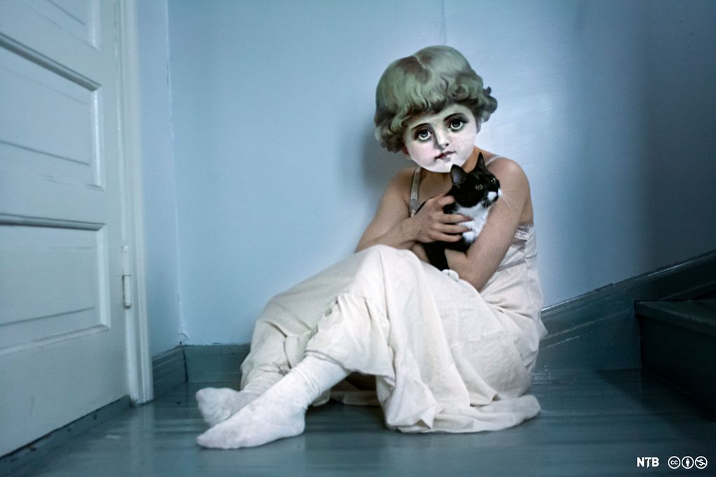 Ei jente med hvit kjole og dukkemaske sitter alene på gulvet og holder en svart og hvit katt. Foto.