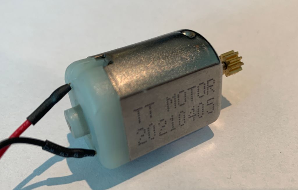DC-motor. En liten firkant i metall og plast, med to ledninger i den ene enden og en tannhjulsforma skrue i motsatt ende. Foto.