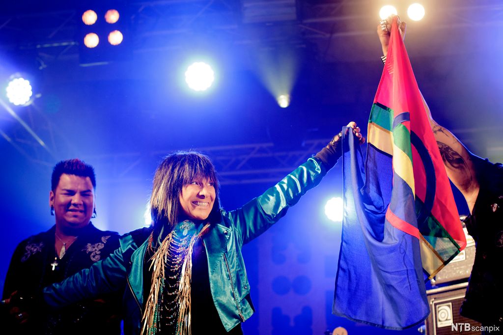 Smilende kvinne på en scene holder et samisk flagg sammen med en person utenfor bildet. Foto.