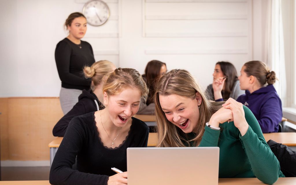 To jenter sitter i et klasserom og ler foran en dataskjerm. Foto.